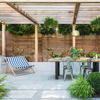 outdoor dining with patio cover ideas, slatted wood pergola with dining table and double deckchair, tiled floor, plants hanging from pergola