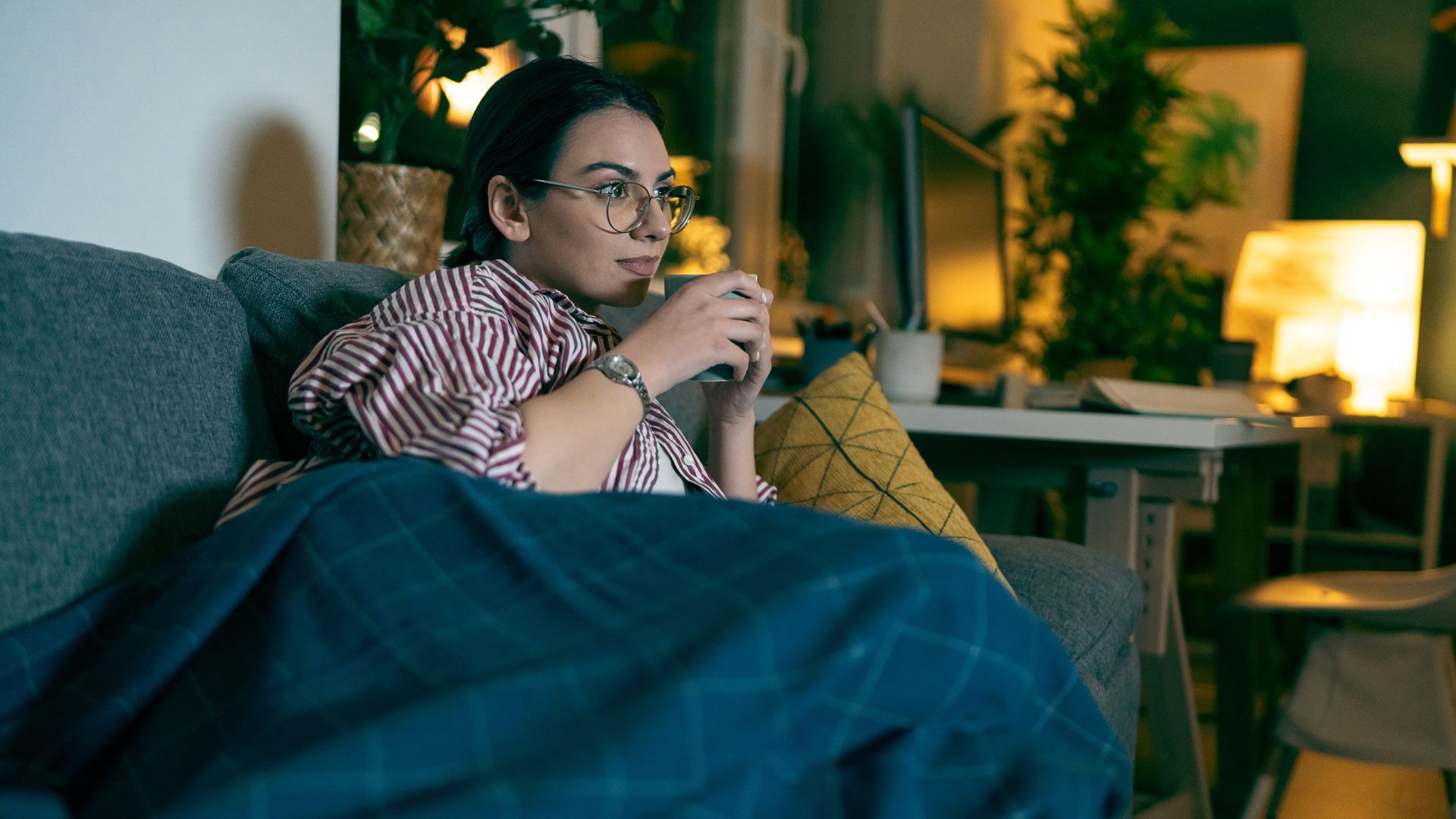 A dark-haired young woman wearing glasses and sipping a mug of tea watches TV on a grey sofa in a dimly-lit living room.