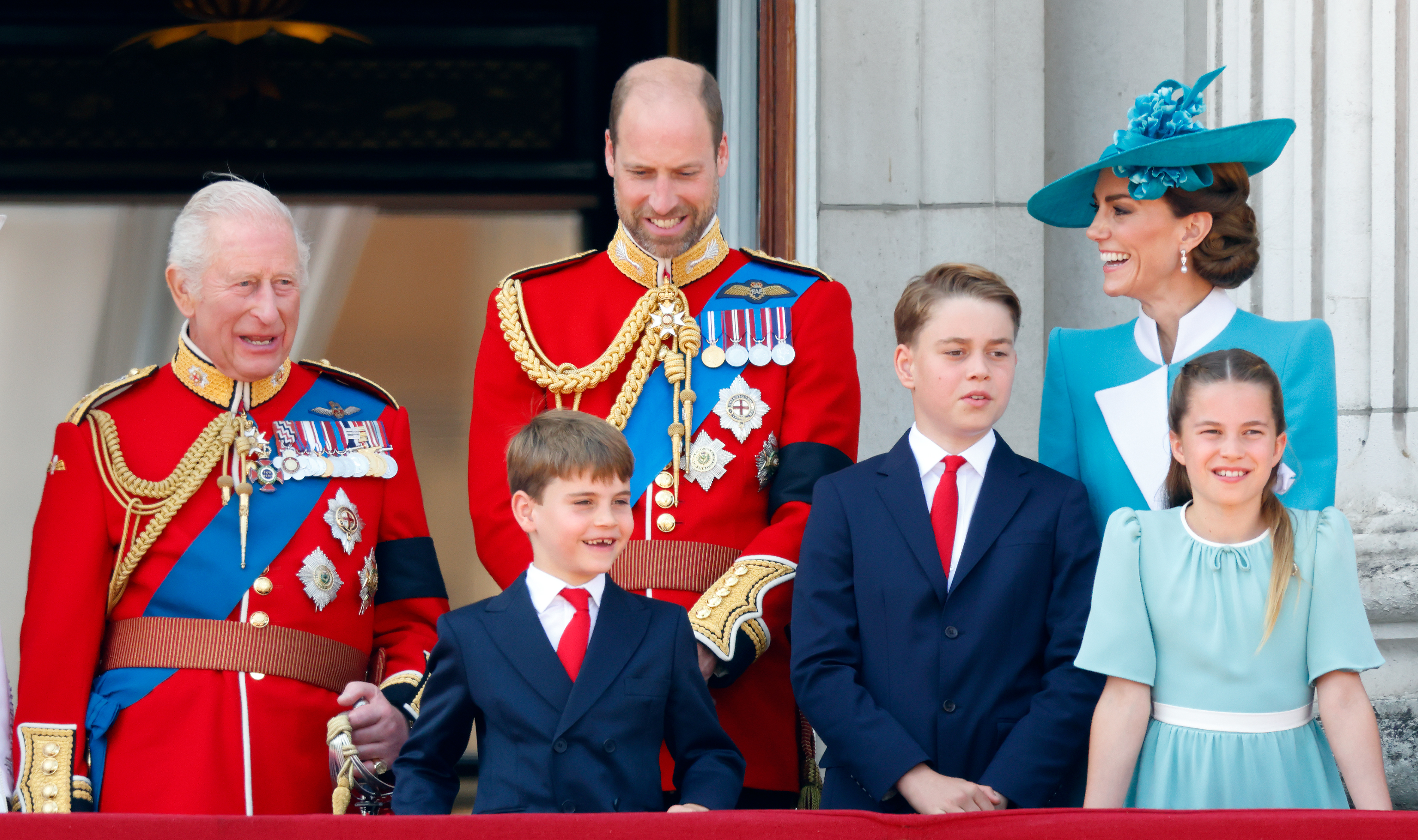 King Charles, Prince William, Prince George, Prince Louis, Princess Charlotte and Princess Kate wearing uniforms and dress outfits on the balcony of Buckingham Palace