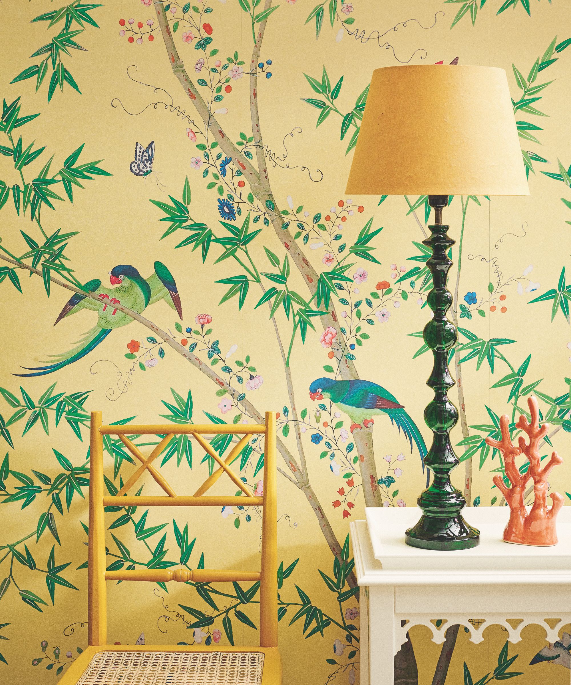 A bright yellow wallpapered room decorated with green trees with birds, a yellow chair, a white table with a green lamp and a decorative orange coral statue.
