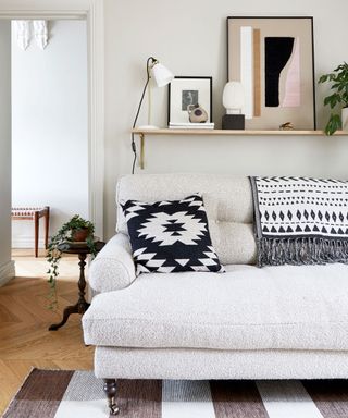 Beige living room ideas illustrated with a roll top sofa, monochrome cushions and a shelf with paintings resting on it.