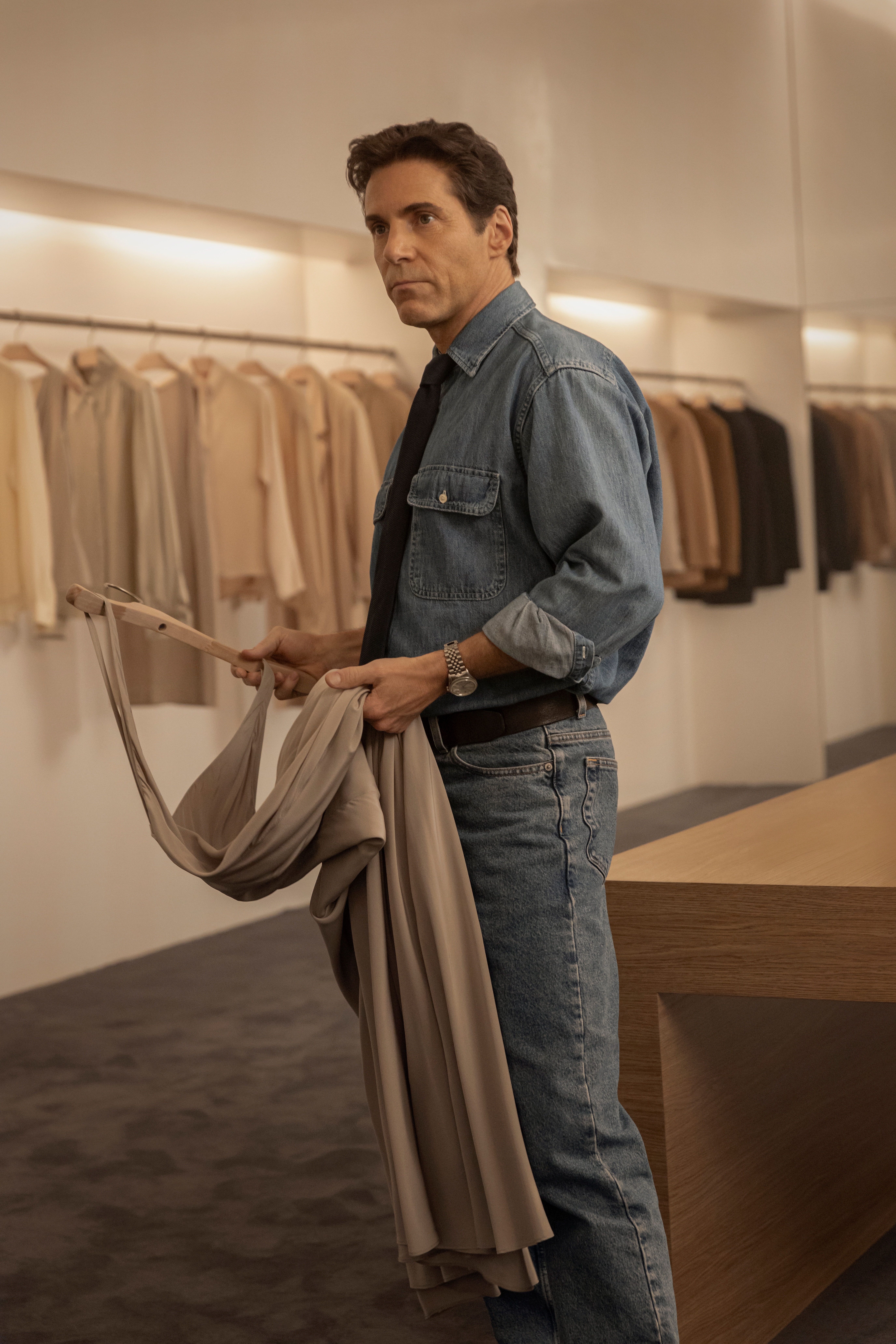 A middle-aged man in a total denim look with a blue tie stands holding a gray maxi dress in a hanger in a dimly lit room with carpeted floors and lots of garments in earthy tones.