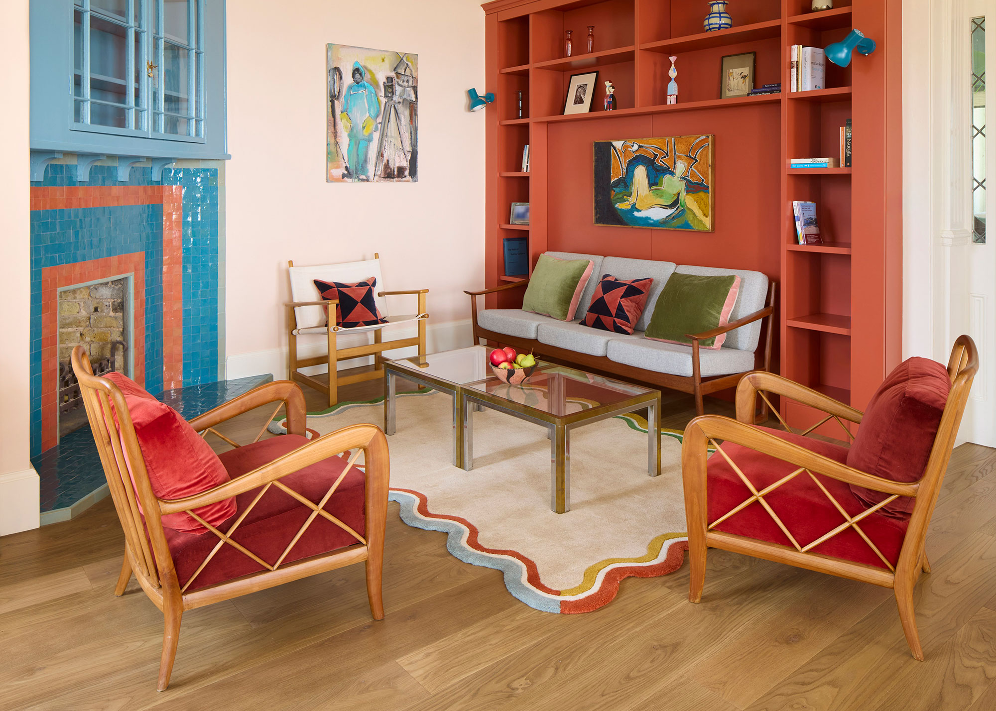 small living room with red painted built-in shelving and blue and orange tiled fireplace with red velvet wooden framed armchairs