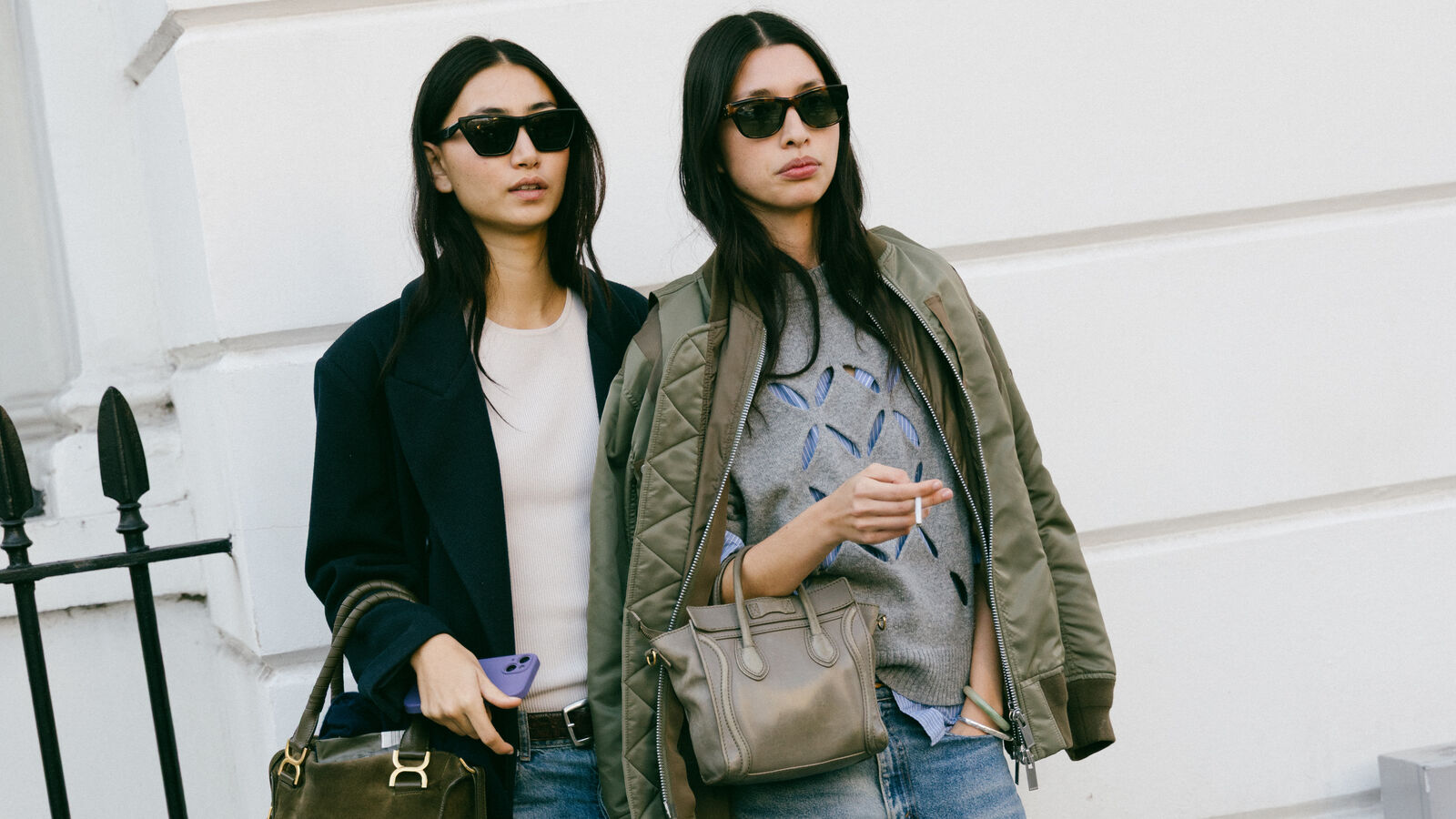 Models pose together for camera. One is wearing a black blazer, tee shirt, and jeans while carrying large handbag.The other friend is wearing a layered gray sweatshirt over jeans with a olive jacket on her shoulders with a asmall handbag. 