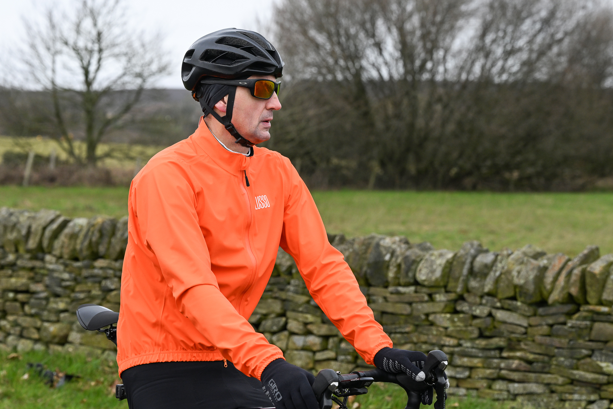 Man wearing an orange jacket, black helmet, black gloves and sunglasses sitting on the top tube of his bike