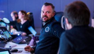 people in dark blue shirts sit at a bank of computers in a mission control room