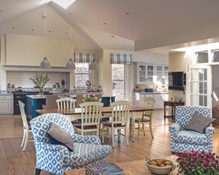 A large kitchen extension with an island below pendant lighting next to a country style dining table and two blue and white patterned armchairs