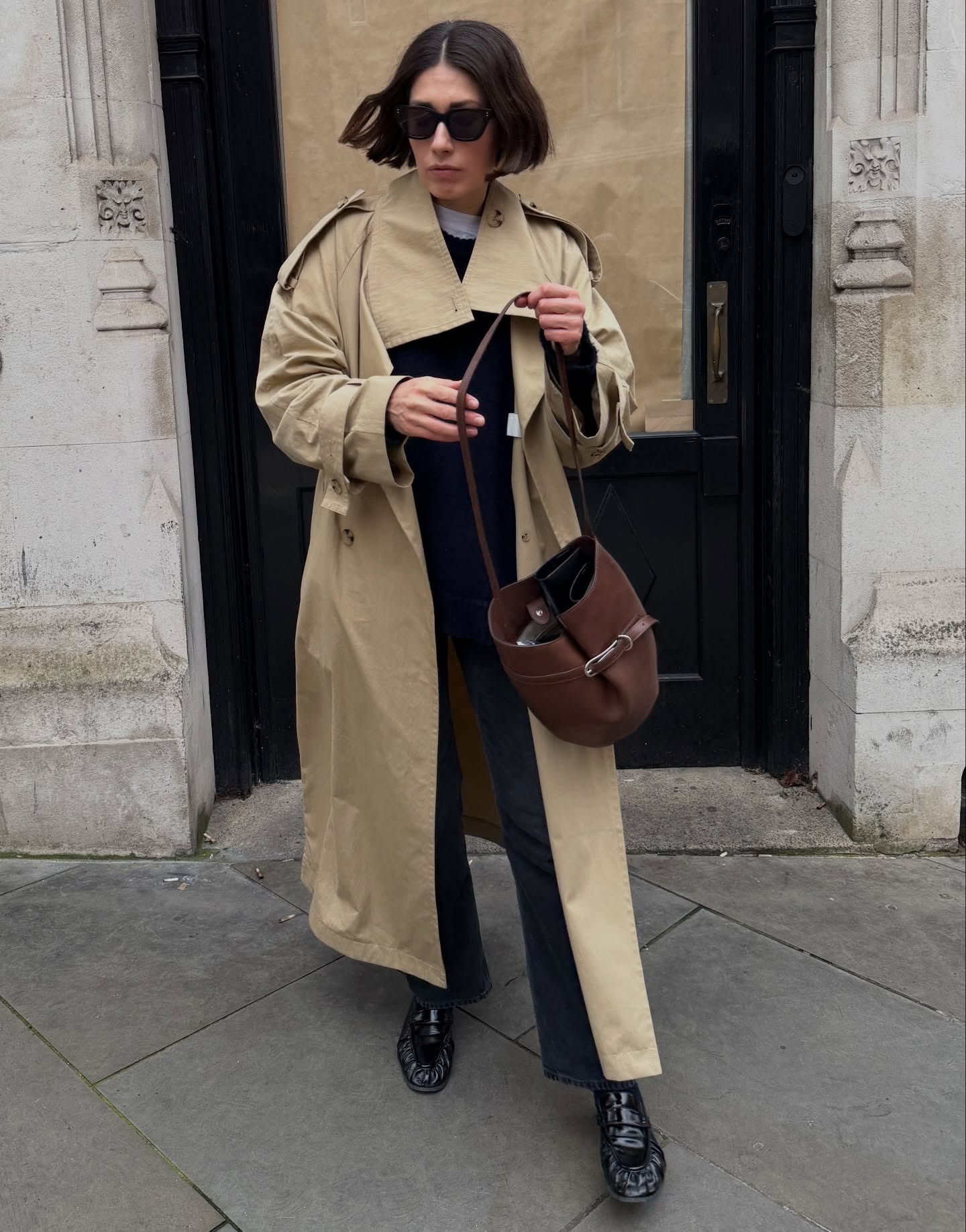 Woman wears beige trench coat, black jeans, black loafers, carries brown Liffner Belted Buckle Bag