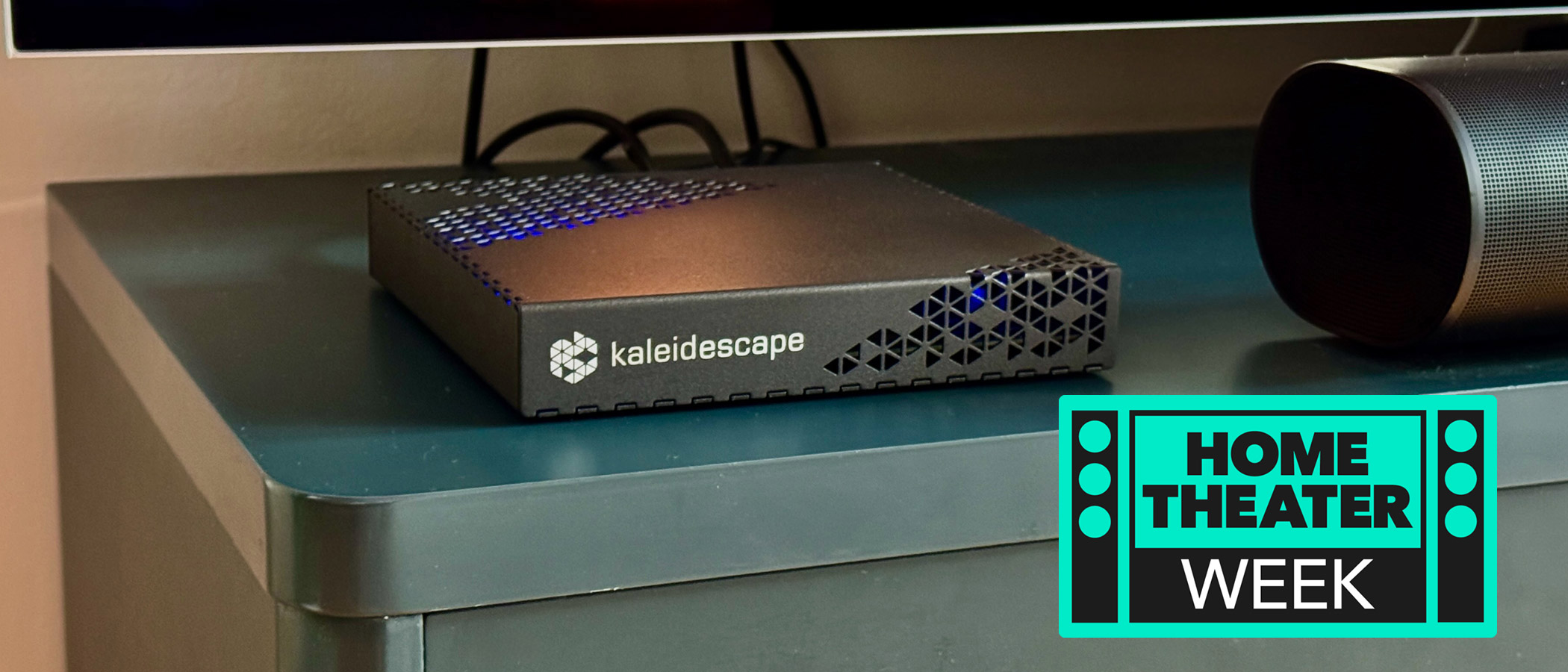 The Kaleidescape Strato E Movie Player on a navy blue TV unit. A badge saying &quot;Home Theater Week&quot; is in the bottom right corner.