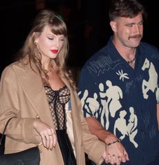 Taylor Swift wearing a tan coat and black bustier holding hands with Travis Kelce, wearing a blue patterned short sleeve shirt and smiling