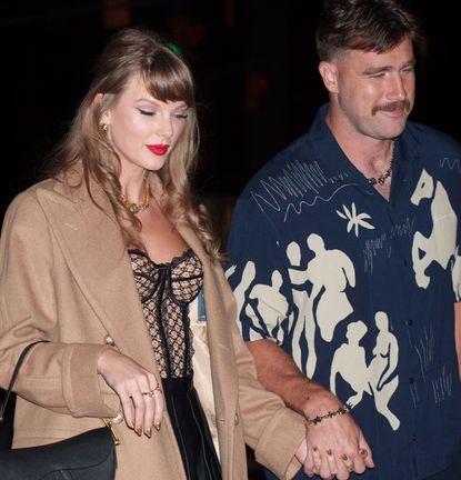 Taylor Swift wearing a tan coat and black bustier holding hands with Travis Kelce, wearing a blue patterned short sleeve shirt and smiling