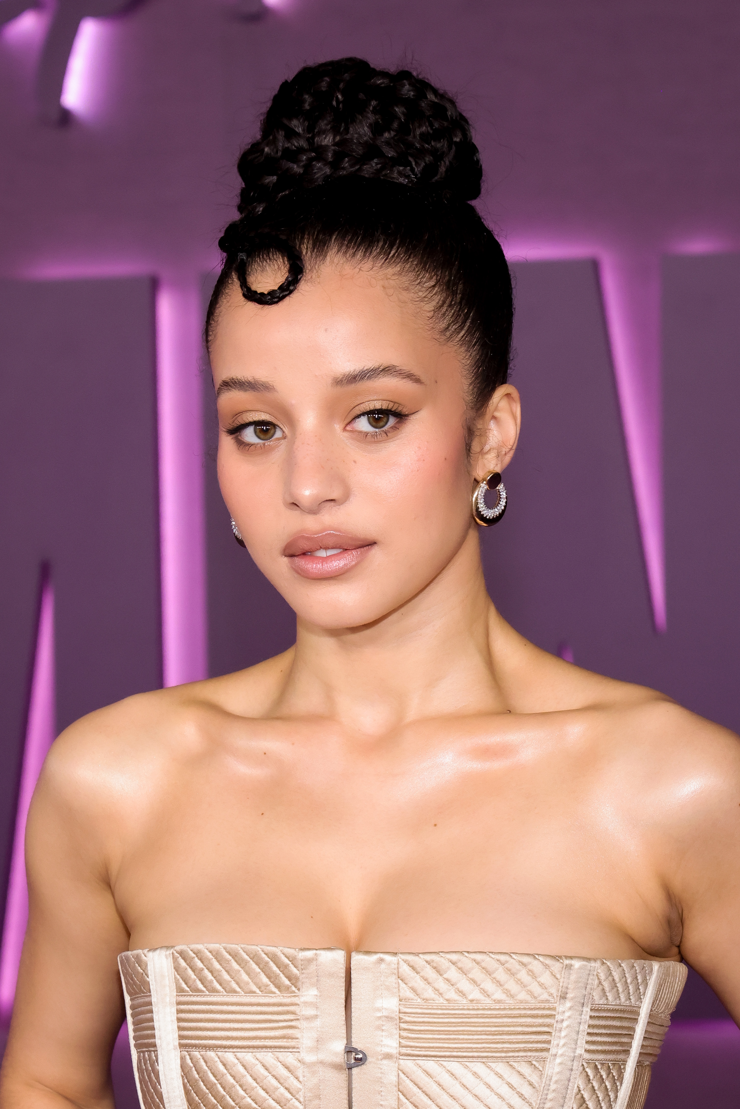 a young woman with dark hair pulled into a high bun wears a strapless, tan dress in front of a purple background