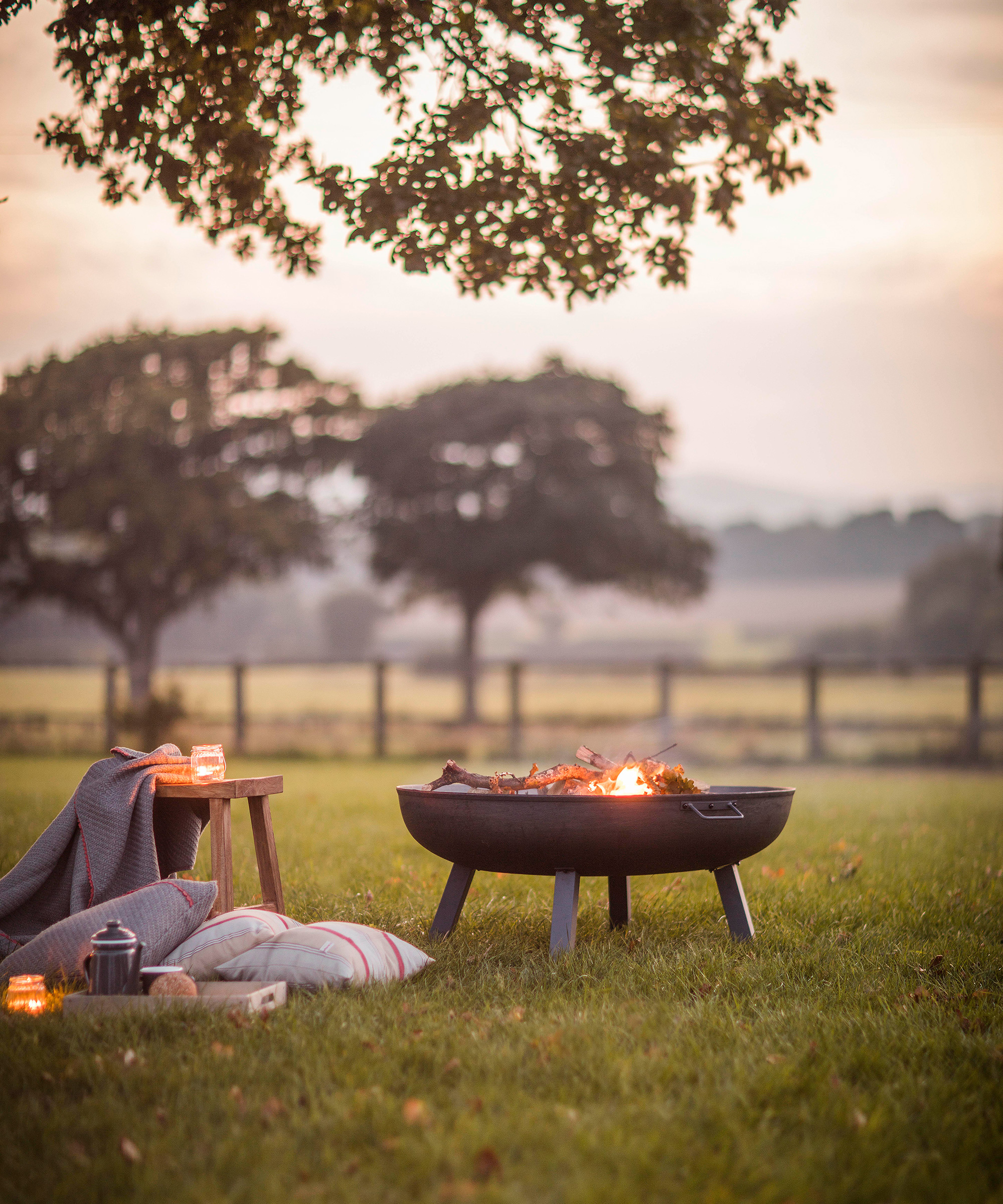 A fire pit on a lawn at sunset with some cushions and a throw