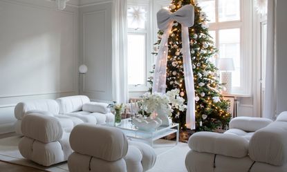 A white living room with white B&B Italia sofa, glass coffee table and Christmas tree with oversized white bow
