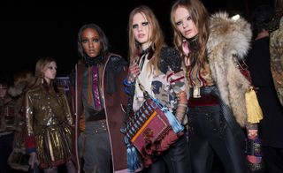 Four female models wearing looks from the Dsquared2 collection. One model is wearing a brown, red and blue jacket and a short dark gold and red skirt. Another model is wearing a grey, purple and orange top, grey trousers and a dark maroon coat with contrasting trims and black detail. The third model is wearing a brown camouflage style top with red and black detail, black trousers and she is holding a maroon, orange and blue bag with tassels. And the fourth model is wearing a red, blue and brown top, belt, black trousers with black detail and a brown sleeveless fur jacket