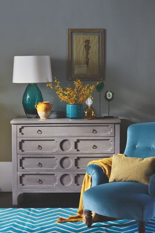 Grey chest of drawers and teal armchair in bedroom with grey walls and teal herringbone rug on floor