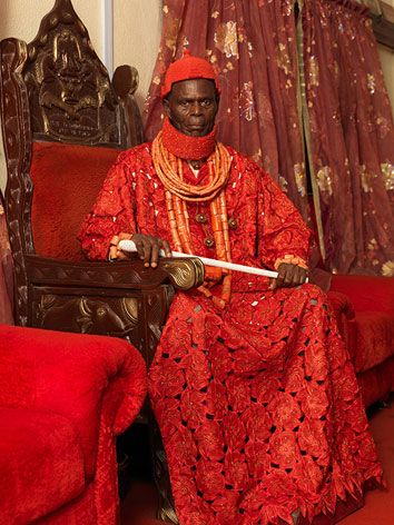 A portrait of an important tribal person sat on a red chair wearing a red/orange robe and a red crown. He is surrounded by two tribal persons holding wood-style objects. On the wall behind are portraits and large wood swords. 