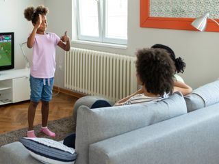 A girl in a pink top and denim shorts playing charades. She is making hand gestures to her parents who are sat on a sofa.