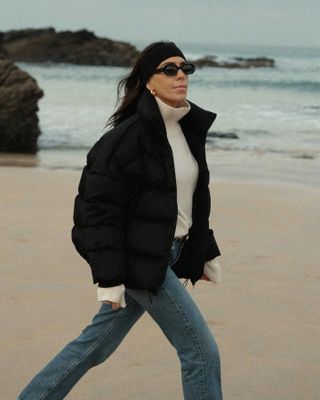 British influencer Jessica Skye walking on a beach in Cornwall wearing oval sunglasses a short black puffer coat, cream funnel neck sweater, and jeans.