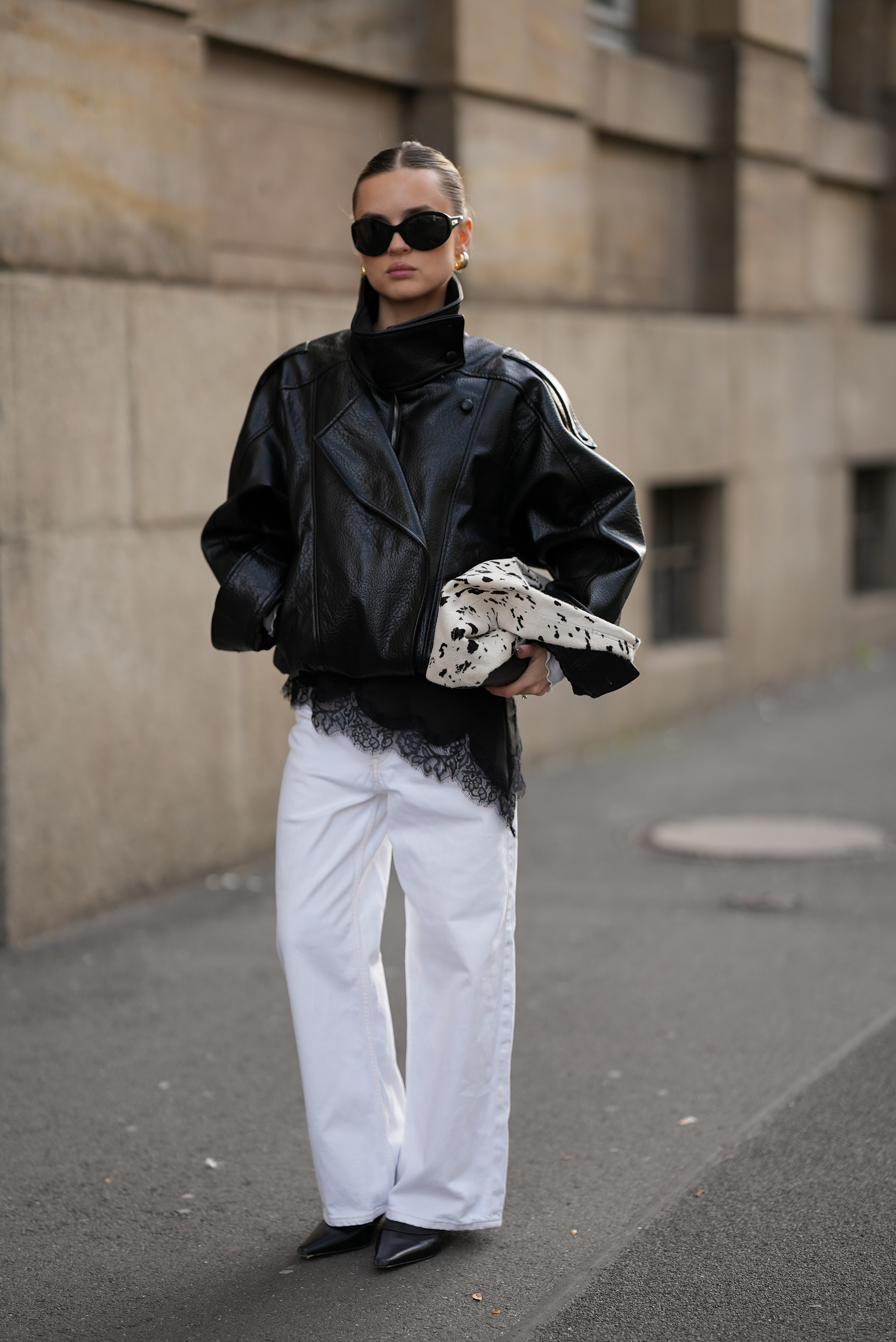Duesseldorf, GERMANY - NOVEMBER 10: Romina Meier is seen wearing an oversized black viral leather PU jacket with a funnel neck collar, wide oversized silhouette and shoulder straps from Zara; a black silk top with asymmetrical lace trim at the hem from Zara; white wide-leg vintage denim pants; a white shoulder bag in pony hair look with bold black speckled pattern; a pair of black patent leather heeled boots with pointed toe from Ance Studios; round black sunglasses from Vogue Eyewear; silver and golden hoop earrings from high by honey hoops, with one in 30mm 18k gold plated and another in 25mm rhodium plated; her blonde hair is styled in a sleek, twisted bun with a center parting on November 10, 2025 in Duesseldorf, Germany. (Photo by Moritz Scholz/Getty Images)