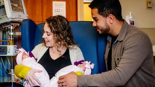 photo of a smiling, white woman with curly hair sitting down and holding two babies swaddled in pink blankets. A tan man with black hair, her husband, is sitting next to them, smiling and reaching over to peer at the children 