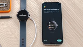 Google Pixel Watch 3 on a wood table next to a Pixel 9 smartphone showing a screen asking users to select which apps they'd like preloaded on their watch during the setup process