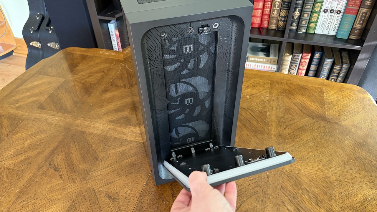 A hand opens the front panel of a Maingear MG-1 MK.II computer tower on a wooden table, revealing fans inside. Books and a guitar case are visible in the background.