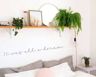 Bright bedroom with wall decal and over bed shelf styled with houseplants, framed picture, and mirror