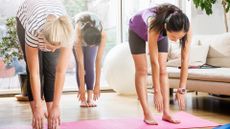 three women in a living room setting performing a roll-down exercise, their heads are down looking at their feet