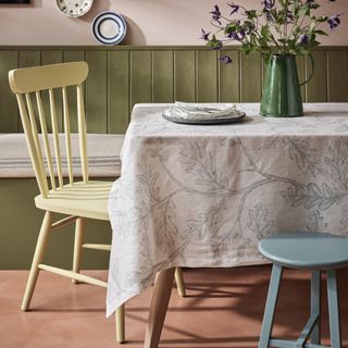 small dining room with bench seating, yellow painted chair, blue painted stool, patterned table cloth, vase of clematis, plates on wall, painted tongue and groove