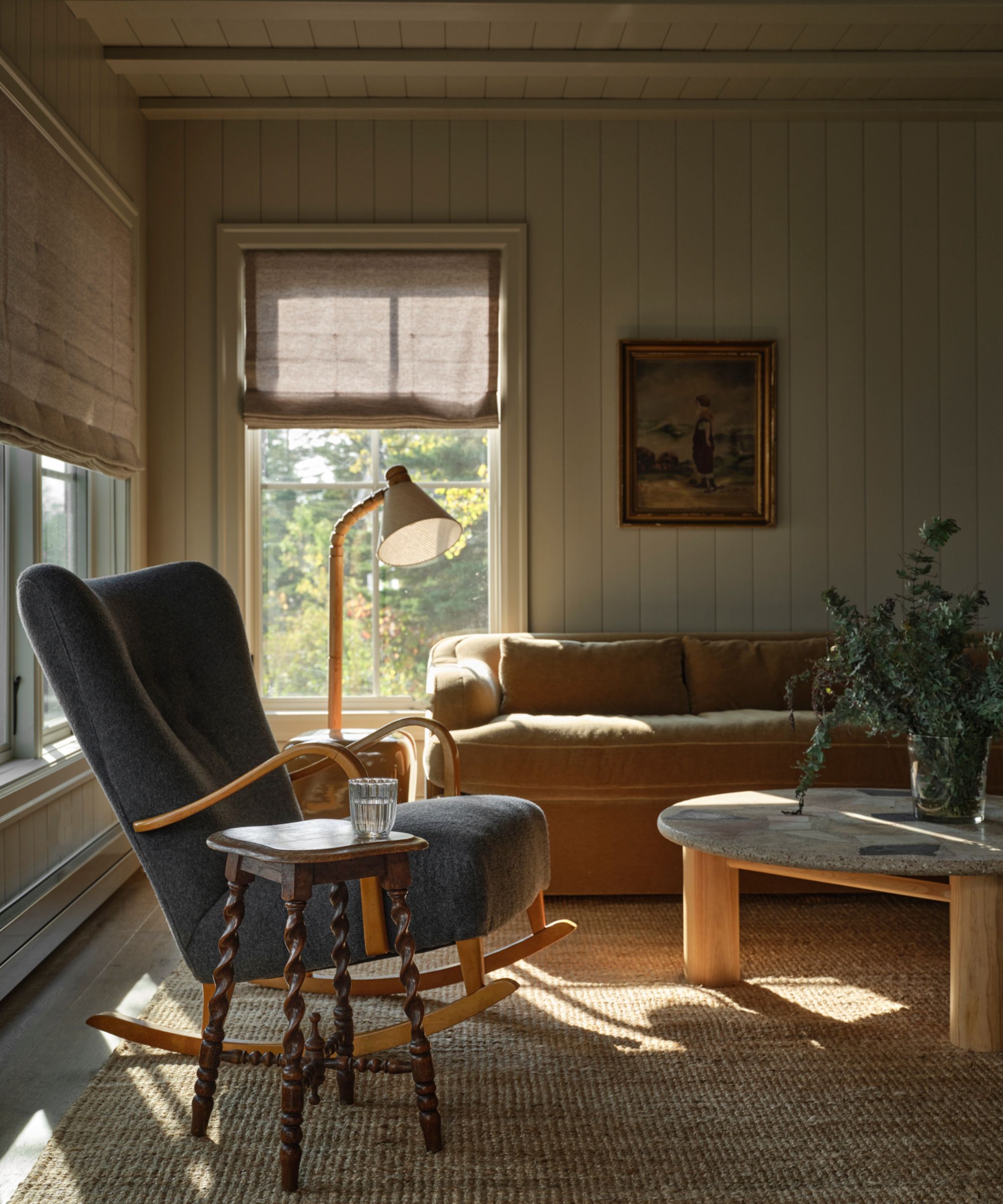 a vintage-styled paneled lakeside cottage living room with a beige velvet couch, vintage stone coffee table and a blue rocking chair