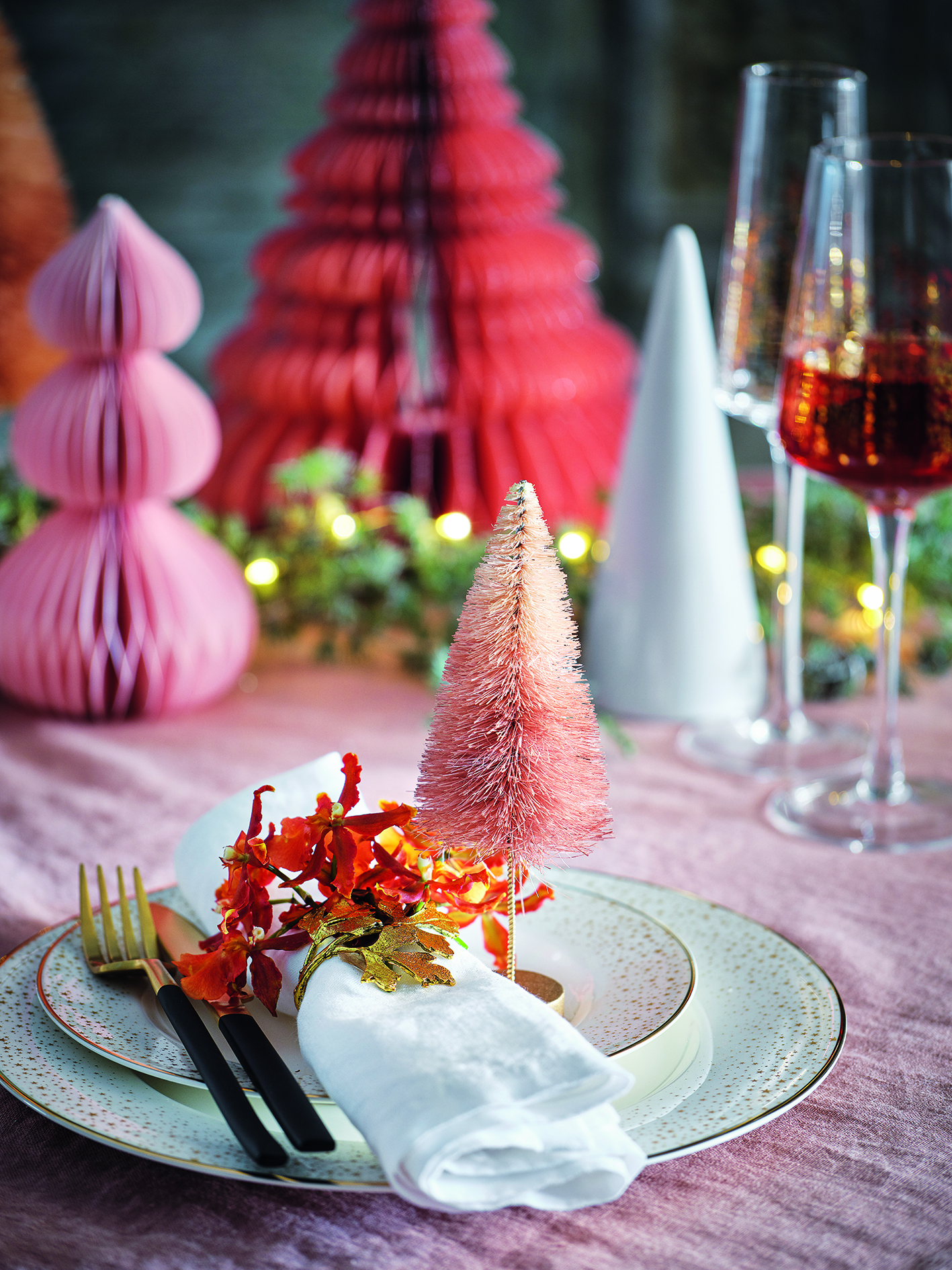 A Christmas table with a dusty pink tablecloth and a dusty pink bottle brush sits on the plate next to a vibrant napkin ring, with paper and ceramic Christmas trees in the background with wine glasses and lit foliage