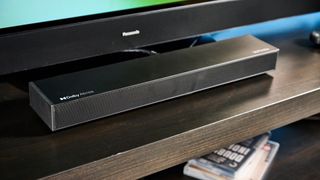 the ultimea nova s50 a black compact soundbar with LED on the front and a wired subwoofer is photographed on a black TV mount connected to a panasonic TV