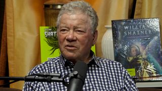 William Shatner talks about selling a kidney stone for charity. He has a microphone in front of him and is wearing a blue and white checkered shirt. 