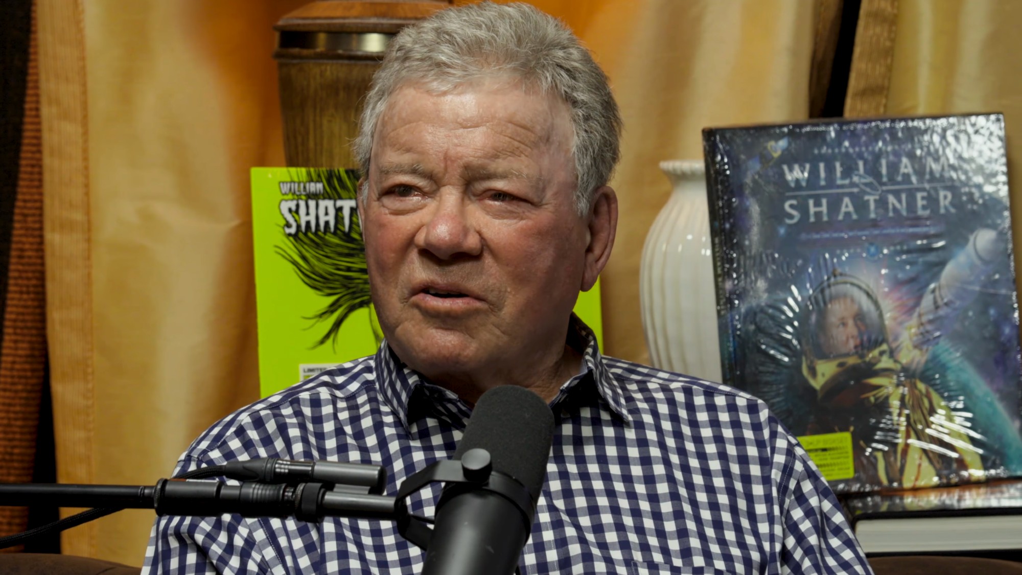 William Shatner talks about selling a kidney stone for charity. He has a microphone in front of him and is wearing a blue and white checkered shirt. 