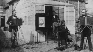 Fox Talbot (1800 -1877) right, is adjusting the lens on a large 'box' camera, whilst an assistant copies an engraving, left