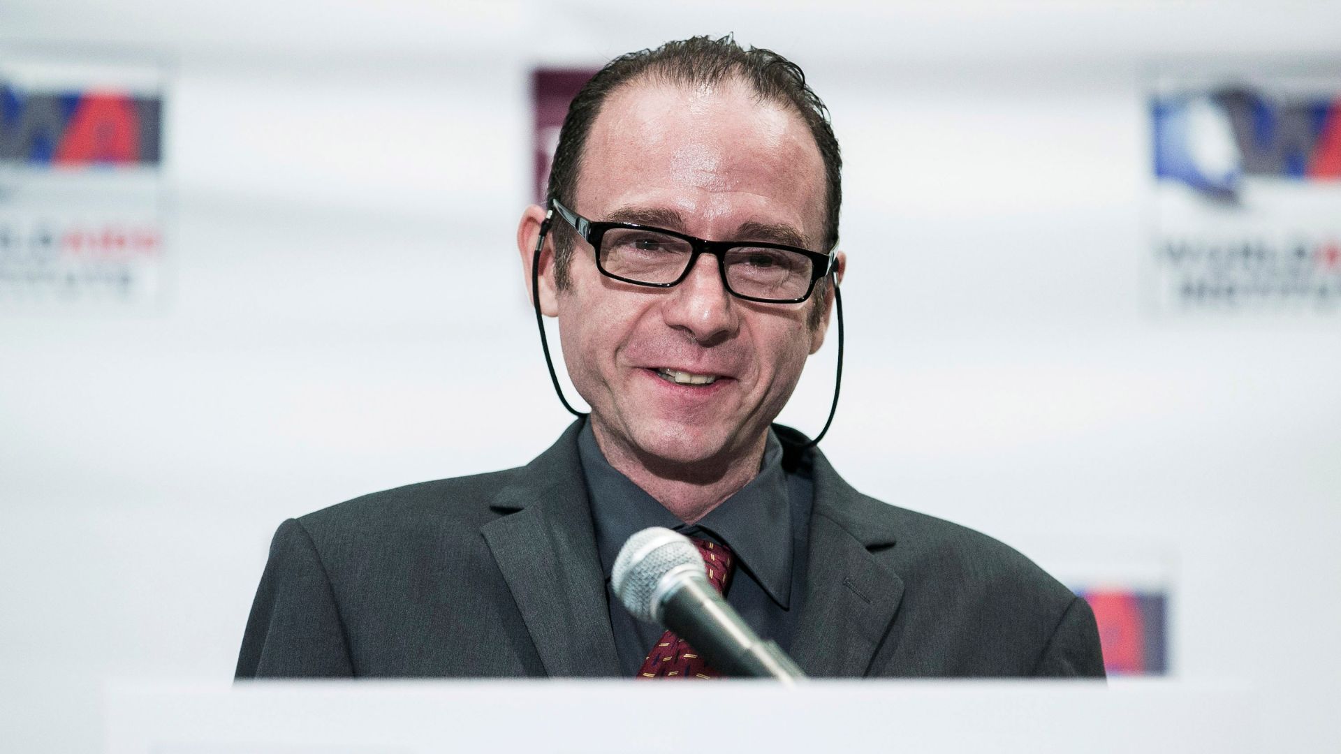 photo shows a white man with short brown hair wearing rimmed glasses and a dark suit as he smiles behind a podium