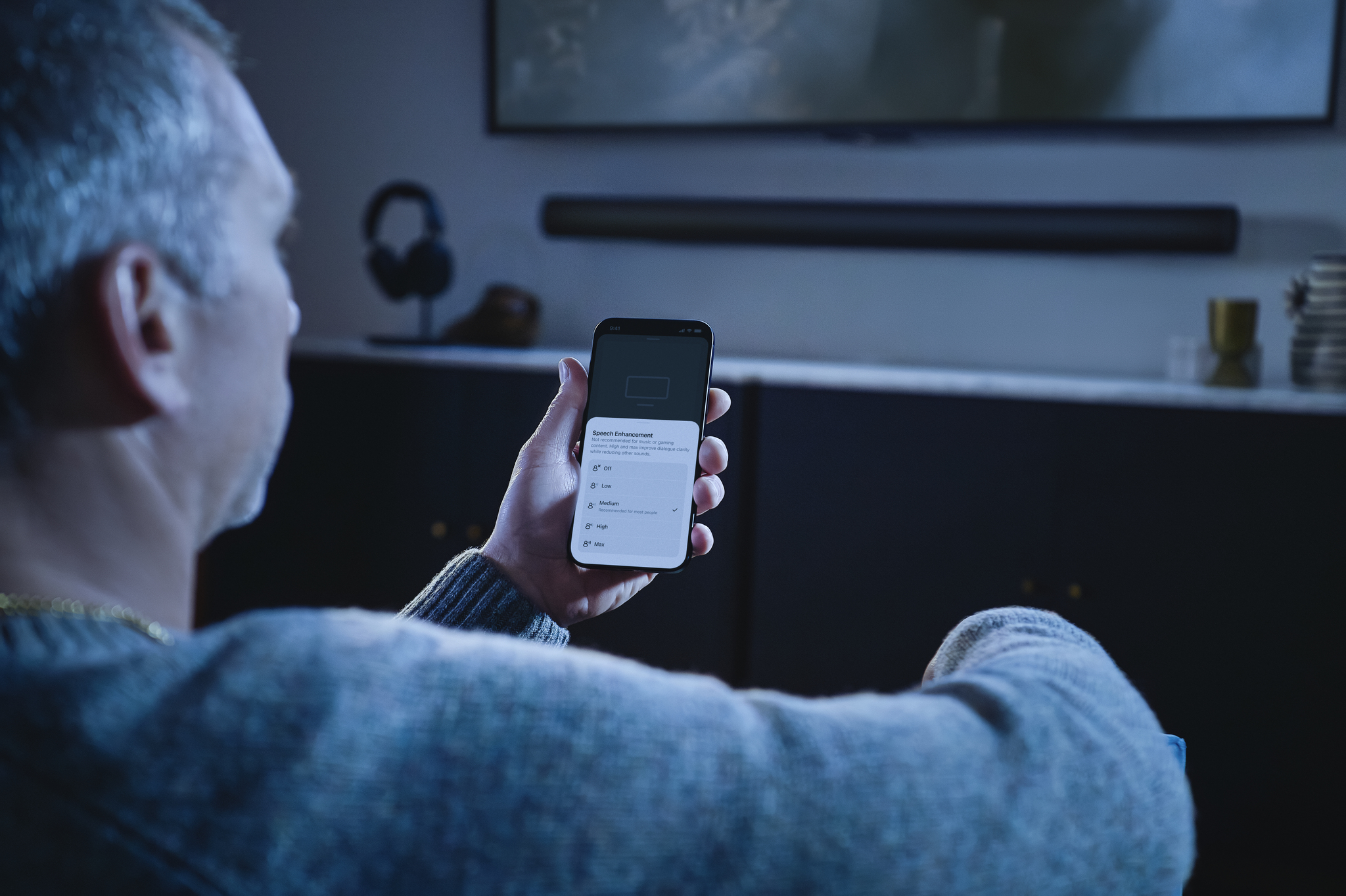 man with grey hair holds his phone in front of him, which is displaying the Sonos Speech Enhancement app. He is in a darkish room and you can see the TV screen slightly in shot above him.