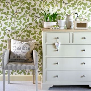 Pale green chest of drawers with grey chair and printed wallpaper