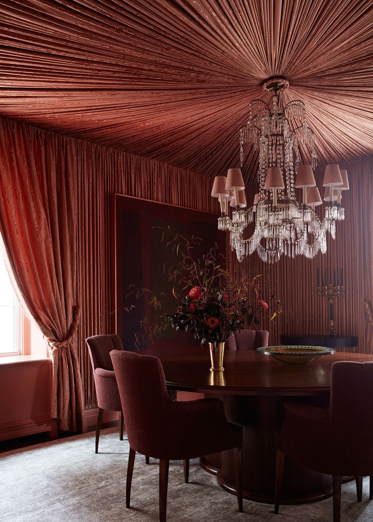 A modern dining room with a tented ceiling in a terracotta color which is continued on to the walls. A large dining table with dark red dining chairs and an opulent chandelier above it.