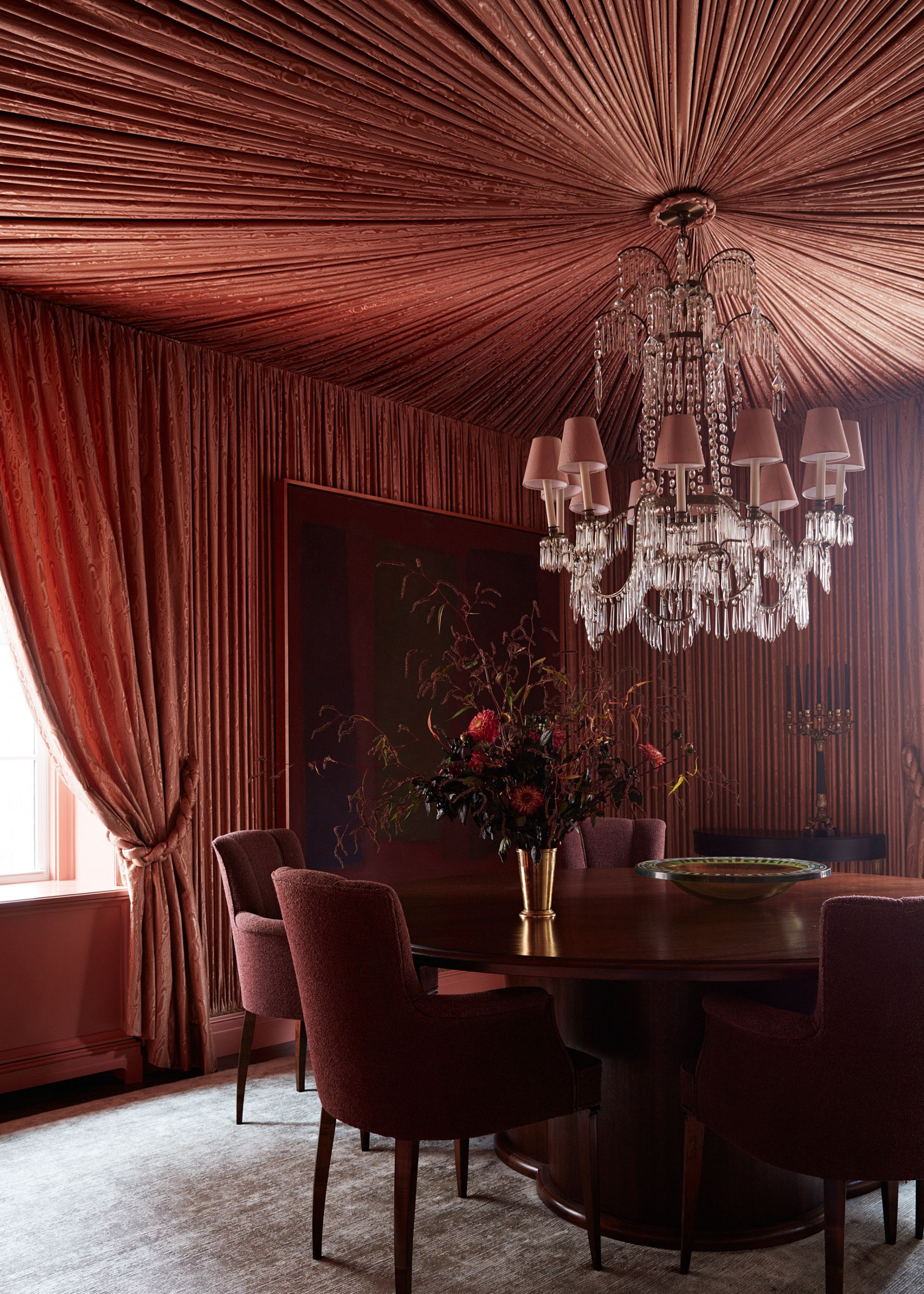 A modern dining room with a tented ceiling in a terracotta color, which is continued on to the walls. A large dining table with dark red dining chairs and an opulent chandelier above it.