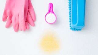 How to clean a mattress from urine image shows a fresh yellow urine stain on a white mattress surrounded by pink rubber cleaning gloves, a pink pot of bicarbonate of soda and a blush scrubbing brush 