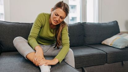 A woman sits on a dark gray couch rubbing her foot and ankle. She wears a green, long-sleeved t-shirt and gray pants.