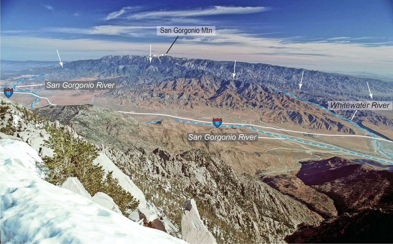 An aerial view of the San Gorgonio pass shows mountains in the foreground, the San Gorgonio river in the middle, and another set of brown wrinkled mountains in the background. White arrows point to the trace of the San Andreas fault through the background mountains.