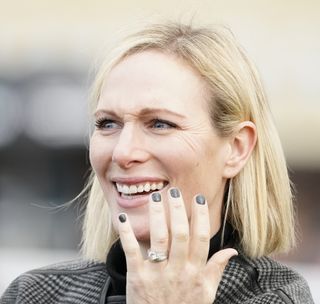 Zara Tindall wearing a black coat and black nail polish holding her hands up to her face laughing