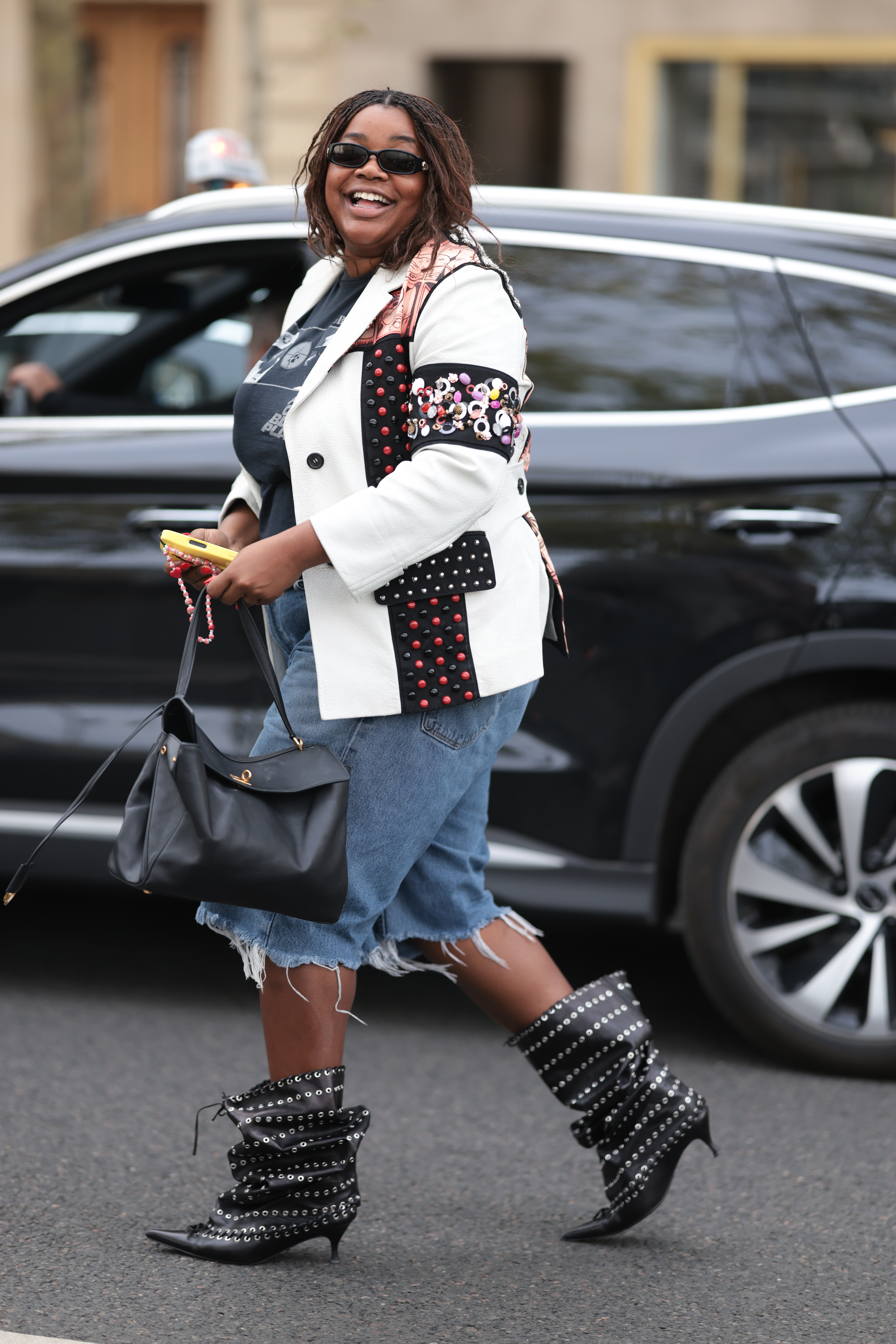 PARIS, FRANCE - OCTOBER 02: Gabriella Karefa-Johnson is seen wearing black sunglasses and has her hair styled in braids. She is dressed in a white blazer featuring black and red stud accents on the sleeves and front, layered over a dark t-shirt. Her cropped denim jeans have distressed hems, and she wears black pointed-toe boots adorned with silver studs. She carries a large black Balenciaga Rodeo handbag over her shoulder during the Rabanne Show Womenswear Spring Summer 2026 as part of Paris Fashion Week on October 02, 2025 in Paris, France. (Photo by Jeremy Moeller/Getty Images)