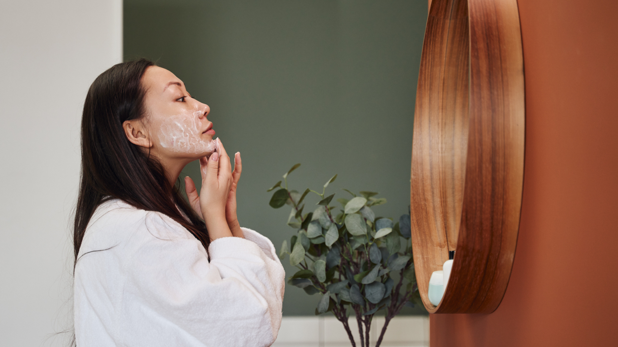 Women in a white robe applying skincare in the mirror