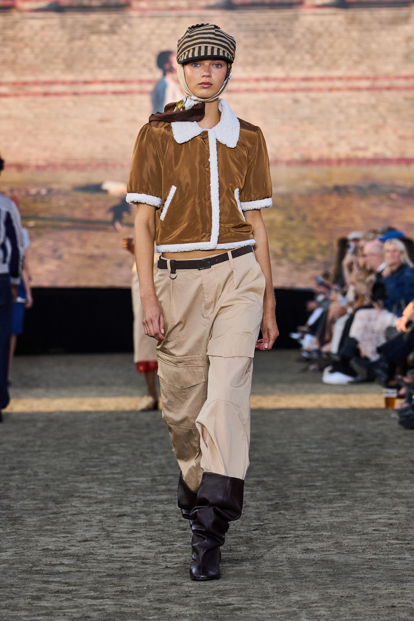 An image from the Baum und Pferdgarten S26 053 Runway show featuring best square-toed boots. The model wears a shearling cardi, tan trousers and a hat. This image was take from Launchmetrics.