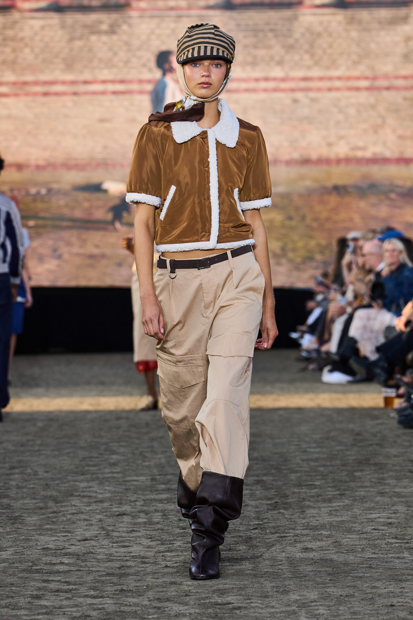 An image from the Baum und Pferdgarten S26 053 Runway show featuring best square-toed boots. The model wears a shearling cardi, tan trousers and a hat. This image was take from Launchmetrics.