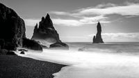Black and white photo of a misty ocean shore with sharp, jagged rock formations rising from the water under a cloudy sky